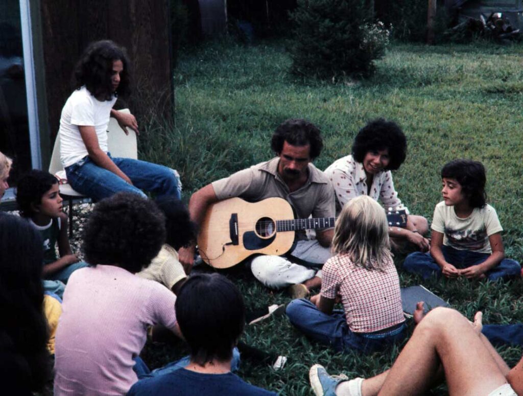 Historic photo of young Appel Farm campers gathered around a guitar instructor, seated on the ground