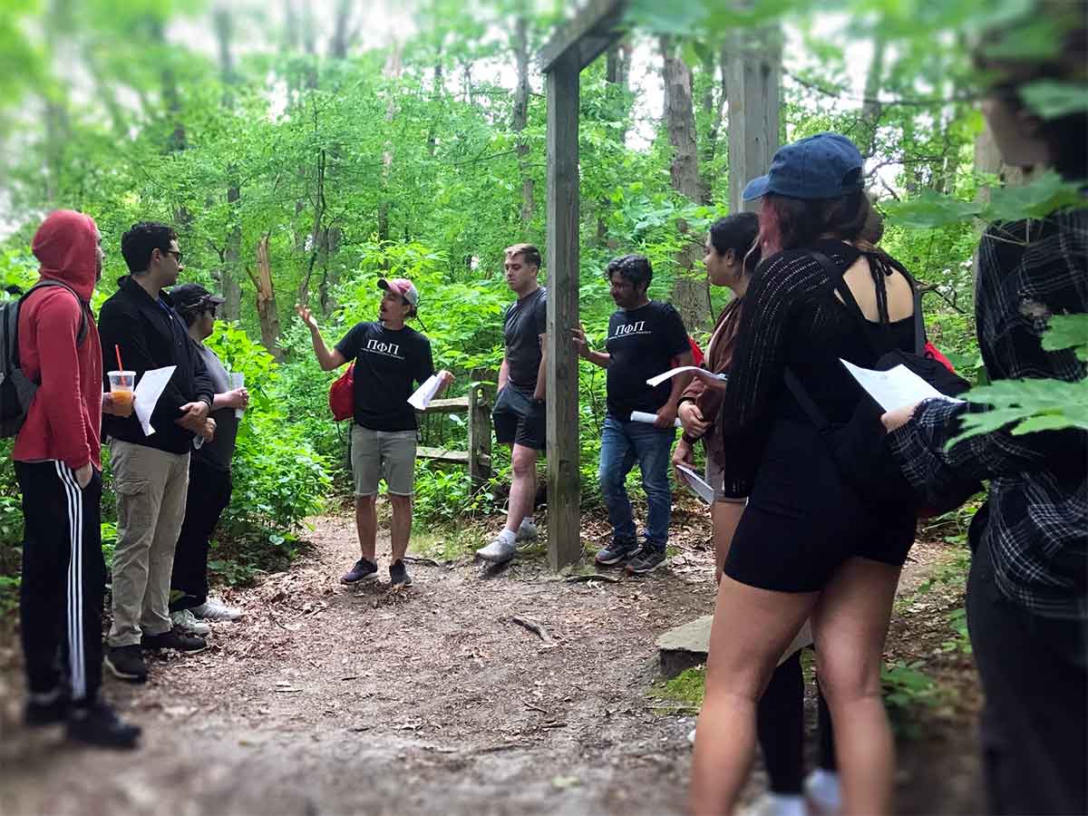 Participants speak during a "Philosophy Walk and Talk" in a green, wooded area underneath a trail entrance sign