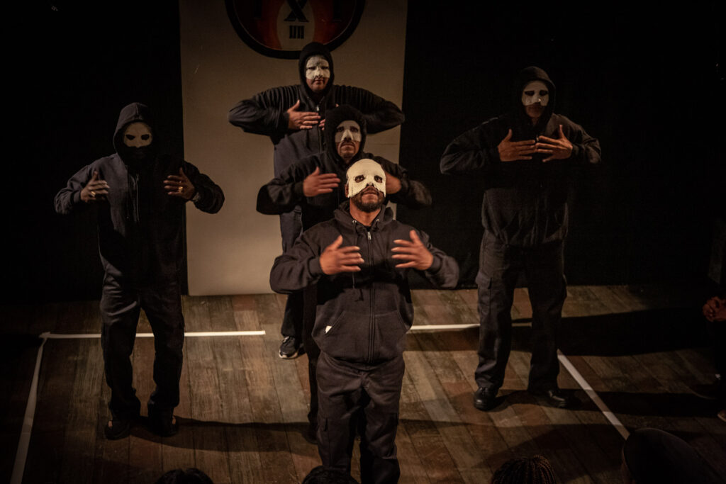 5 Returning Citizens dressed in black with white "stigma" masks perform on stage