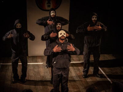 5 Returning Citizens dressed in black with white "stigma" masks perform on stage