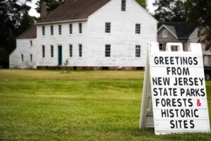 A sandwich board sign reading "Greetings from New Jersey State Parks Forests & Historic Sites" in the foreground with a white building in the background