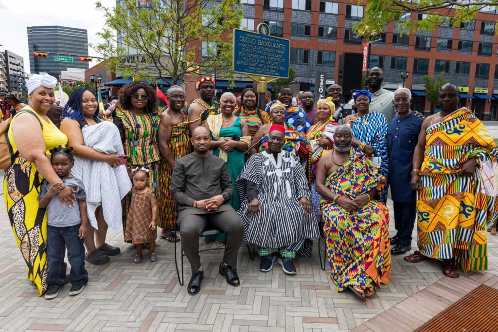 Group of about two dozen people, some seated, some standing, in front of the Cudjo Banquante historical marker outside of NJPAC.