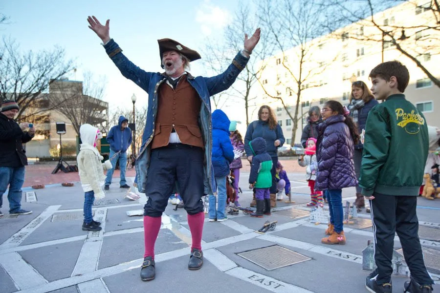 A man in Revolutionary-Era historic clothing speaks on a crowded town square